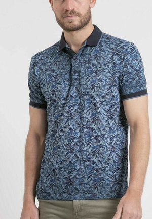 Homme portant un polo bleu à motifs floraux avec un col foncé et des poignets de manches foncés, debout devant un fond clair uni.
