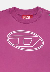 Ljubičasti dukserica s istaknutim bijelim Diesel logom u ovalnom obliku. Ovratnik ima rebrasti detalj i metalne kopče za zatvaranje.