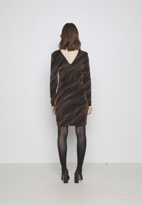 Schwarzes Kleid mit V-Ausschnitt, das ein braunes und goldenes abstraktes Muster aufweist. Lange Ärmel, figurbetonte Passform, getragen mit schwarzen Strumpfhosen und Absatzschuhen.