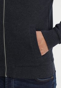 Sweat à capuche zippé gris foncé avec ourlet et poignets côtelés, poches latérales et détail de fermeture éclair argentée. Texture de tissu douce visible.