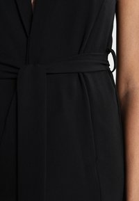 Schwarzes Kleid aus glattem Stoff, mit einem Wickeldesign und einem gebundenen Gürtel. Das Detail zeigt den Taillenbereich und eine Nahaufnahme der Textur.