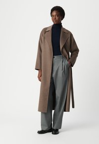 Cappotto lungo marrone con colletto a rever, abbinato a dolcevita blu e pantaloni grigi plissettati. Tessuto morbido, vestibilità comoda e tasche laterali.