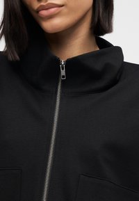 Sort lynlåsjakke med høj krave, glat stof og metallynlåsdetaljer. To horisontale lommer er synlige foran.