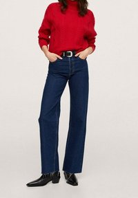 Pull rouge en maille avec motifs en losange, col montant, associé à un jean large taille haute bleu et des bottines noires à talons. Ceinture noire incluse.