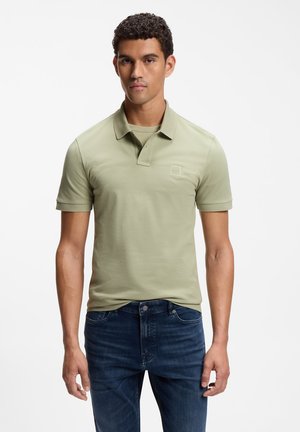 Ung mand med kort krøllet hår iført en lysegrøn poloshirt og mørkeblå jeans står mod en hvid baggrund.