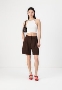 Hvit, kort topp, brune, plisserte shorts og røde høye sko. Liten svart håndveske med skulderstropp. Enkelt bakgrunn.