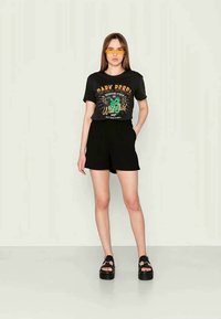 Svart grafisk t-shirt med grön och orange design, tillsammans med svarta shorts. Modellen bär gula solglasögon och svarta plattformssandaler.