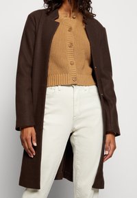 Manteau long marron en tissu lisse, porté sur un cardigan beige en maille avec des boutons. Un jean blanc complète la tenue.