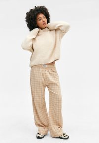 Beige Strickpullover mit hohem Kragen, kombiniert mit weiten, karierten beigen und cremefarbenen Karohosen, die Knöpfe am Bund haben.