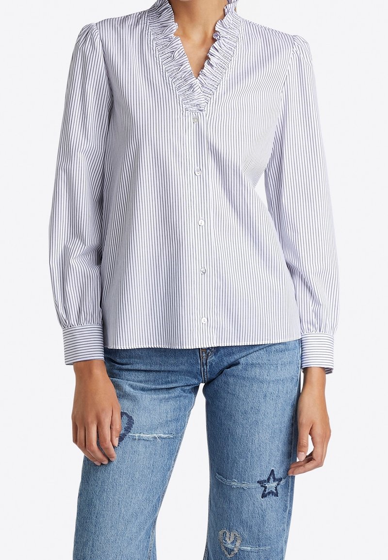 Weiße Bluse mit marineblauen vertikalen Streifen, Rüschenausschnitt in V-Form, langen puffärmeligen Ärmeln und Knopfverschluss, kombiniert mit verwaschenen blauen Jeans.