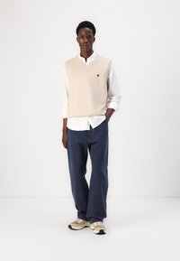 Μπεζ πλεχτό γιλέκο πάνω από ένα λευκό πουκάμισο, συνδυασμένο με σκούρα μπλε φαρδιά παντελόνια και μπεζ αθλητικά παπούτσια. Περιλαμβάνει ένα μικρό λογότυπο στο γιλέκο.