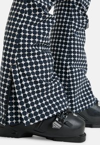 Czarno-białe spodnie narciarskie w groszki z zamkami błyskawicznymi przy kostkach, w połączeniu z czarnymi butami narciarskimi z metalicznymi akcentami i teksturowanymi powierzchniami.