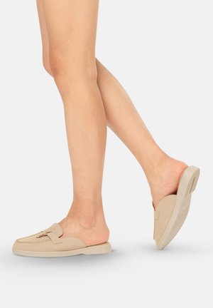 Ben met beige slip-on loafers met gouden gespdetail, één voet plat en de andere omhoog, tegen een effen lichte achtergrond.