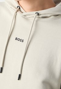 Světle béžová mikina vyrobená z měkké tkaniny, která má klokaní kapsu, stahovací šňůrky a černé logo "BOSS" na hrudi.
