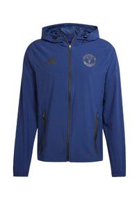 Blå zip-jakke med hætte, med sorte detaljer og Manchester United-logo på brystet. Lavet af åndbart stof med sidelommer.
