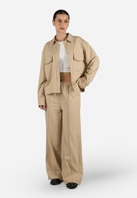 Giacca beige oversize con due tasche frontali, abbinata a pantaloni larghi. Il materiale è liscio; sopra c'è una maglietta bianca a trama corta. Calzature nere.