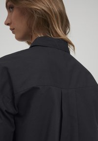Black cotton shirt featuring a classic collar, back pleat detail, and smooth texture. Side seams are clean with no visible hardware.