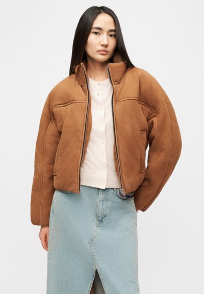 Veste bomber en daim marron avec un col montant, une fermeture éclair à l'avant et des poches latérales, portée par-dessus un haut beige clair et une jupe en denim.