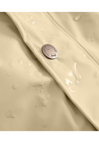 Beige waterdichte jasstof met waterdruppels op het oppervlak, met een zilveren metalen knoop met een gegraveerd logo. Glad textuur.