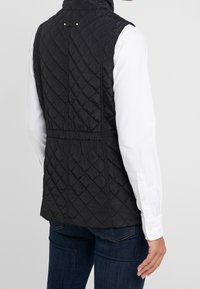 Person som bär svart vadderad ärmlös väst över vit långärmad skjorta och mörkblå jeans, visad bakifrån mot enkel bakgrund.