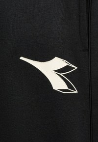 I pantaloni della tuta neri presentano un design grafico bianco prominente che assomiglia a una lettera stilizzata sulla coscia, con una texture liscia e una cintura elastica.