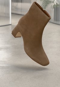 Bota de tornozelo de camurça castanha com salto blocado baixo e zipper lateral sobre um piso cinza claro com fundo de painéis verticais.