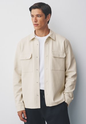 Fiatal férfi rövid, sötét hajjal, bézs színű gombos dzsekit visel fehér ing fölött, egy egyszerű világosszürke háttér előtt áll.