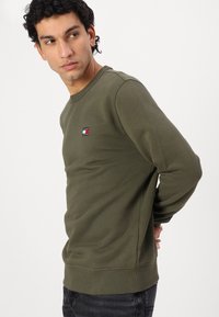 Felpa verde oliva realizzata in tessuto texturizzato, dotata di un collo rotondo e di un piccolo patch con il logo Tommy Jeans sul petto.