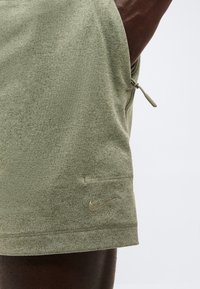 Κοντινό πλάνο ενός ανοιχτοπράσινου υφασμάτινου ενδύματος με πλευρικό φερμουάρ και ραμμένο λογότυπο Nike κοντά στο στρίφωμα, στο μηρό ενός ατόμου.