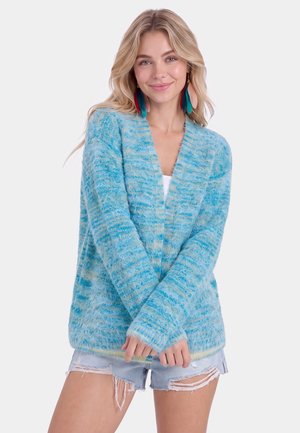 Jonge vrouw lacht, draagt grote meerkleurige veeroorbellen, een blauwe zachte cardigan en gescheurde denim shorts, staand tegen een effen achtergrond.