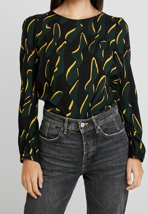 Femme portant un chemisier noir à manches longues avec des formes abstraites vertes et jaunes, rentré dans un jean noir délavé taille haute.