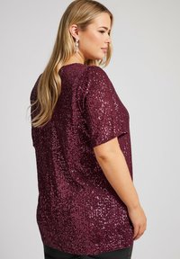 Kimalainen luumunvärinen paita lyhyillä hihoilla, jossa on täysi paljettipinta ja rento malli, nähtynä sivulta.