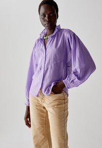 Camicia lavanda con bottoni e maniche a sbuffo, realizzata in tessuto leggero, abbinata a pantaloni beige. Presenta una vestibilità rilassata e linee di cucitura sottili.