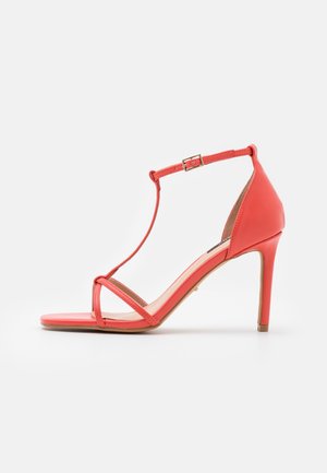 Korale sandały na wysokim obcasie z paskiem T, z cienkimi, łukowatymi paskami, wykonane z materiału syntetycznego, z ozdobnym złotym zapięciem.