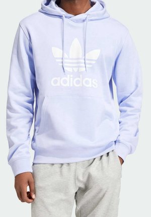 Mężczyzna w jasno fioletowej bluzie z kapturem Adidas z białym logo i jasnoszarych spodniach dresowych, stoi z jedną ręką w kieszeni, a drugą opuszczoną.