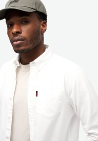 Camisa blanca de botones con un bolsillo y una pequeña etiqueta de marca, superpuesta a una camiseta gris claro. Se lleva una gorra gris en la cabeza.