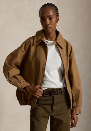 Femme aux cheveux tressés portant une veste marron à fermeture éclair, un haut blanc côtelé, un pantalon marron foncé et une ceinture sombre, debout devant un fond uni.