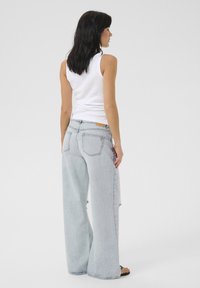 Jeans de pierna ancha de color azul claro con un dobladillo deshilachado, combinados con una camiseta sin mangas blanca ajustada. Presentan bolsillos y una etiqueta beige sobre el bolsillo trasero derecho.