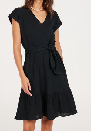 Femme portant une robe noire à manches courtes et col en V, avec une ceinture à la taille et un ourlet à volants, accessoirisée d'un collier de perles.