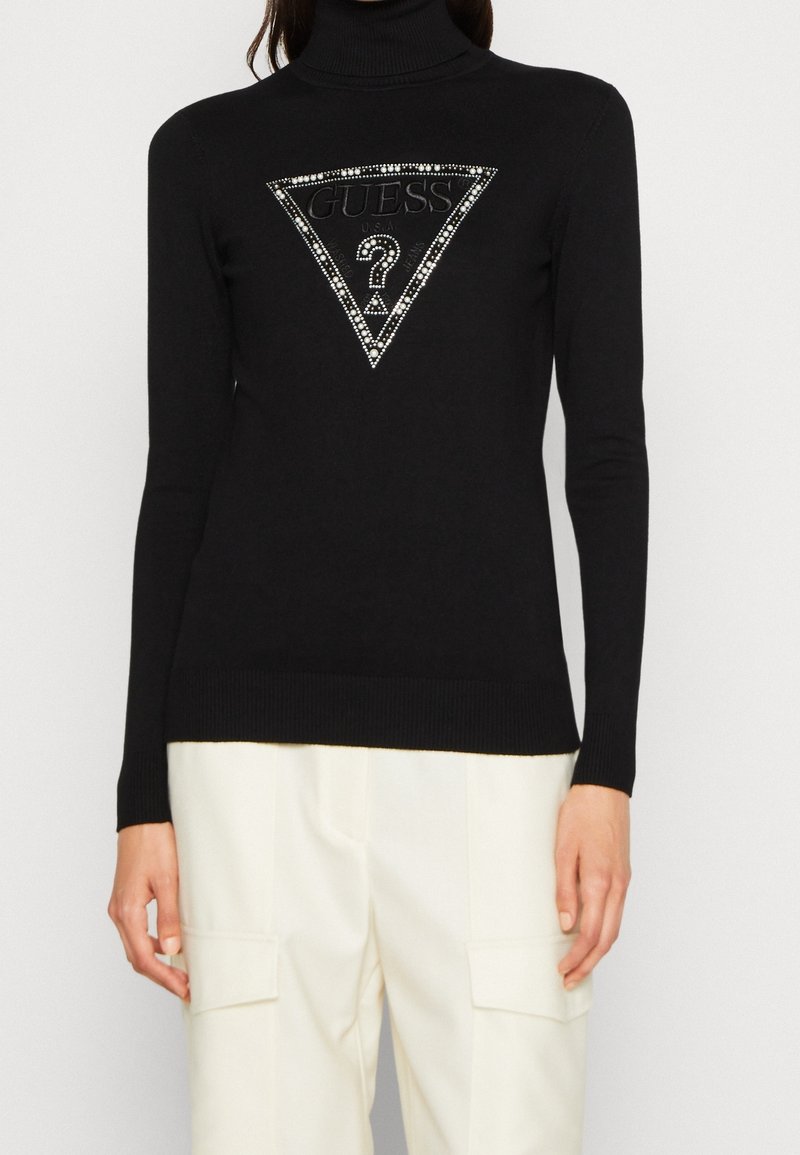 Pull à col roulé noir avec un triangle logo Guess orné de strass sur la poitrine, assorti à un pantalon cargo blanc.