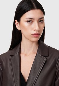 Blazer in pelle marrone con colletto strutturato, design monopetto e texture liscia. Il modello indossa una maglietta nera e orecchini a cerchio dorati.