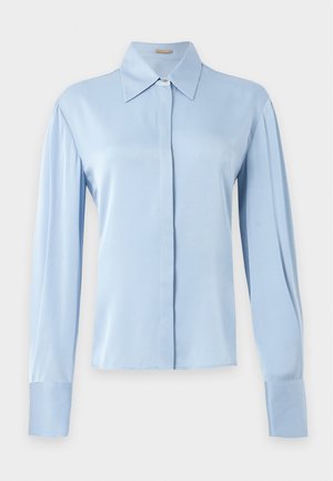 Femeie purtând o bluză bleu cu mâneci lungi și pantaloni largi bej, stând cu o mână în buzunar și sandale negre cu toc.