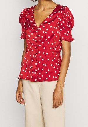 Rød bluse med korte ærmer med hvide prikker, knaplukning foran, puffede skuldre, båret med beige bukser.