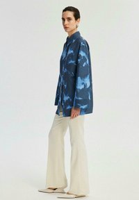 Camicia button-up a righe blu tie-dye con maniche lunghe, abbinata a pantaloni a campana crema e ballerine beige a punta. Tessuto morbido e vestibilità comoda.