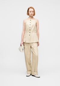 Γυναίκα με κόκκινα μαλλιά που φοράει μπεζ παντελόνι με φαρδιά μπατζάκια και αμάνικο τοπ με κουμπιά, κρατώντας μια μικρή μπεζ τσάντα, στέκεται σε λευκό φόντο.