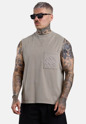 Mann mit kurzen Haaren und Nacken-Tattoo, der schwarze Sonnenbrillen und ein ärmelloses beiges Hemd mit besticktem Taschendesign trägt.