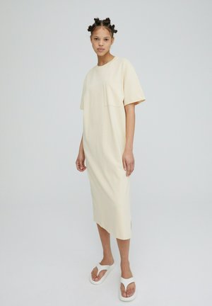 Cream-colored oversized T-shirt dress with a pocket, short sleeves, and side slits, paired with white slip-on sandals. Smooth fabric texture.