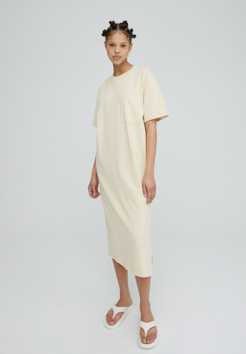 Crème-kleurige oversized T-shirt jurk met een zak, korte mouwen en zijsplitten, gecombineerd met witte instapsandalen. Soepele stof textuur.