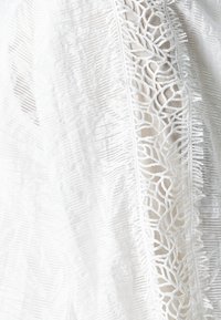 Vitt broderat tyg med bladmönster, med fransade kanter och en texturerad yta som skapar visuell djup och intrikata detaljer.