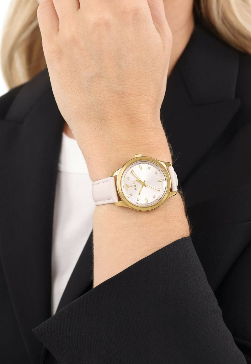 Goldfarbene Armbanduhr mit einem weißen Zifferblatt, goldenen Stundeneinteilungen und einem hellrosa Lederarmband, getragen an einer Hand gegen ein schwarzes Blazer.